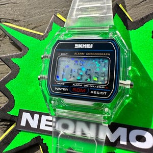 May include: A digital wristwatch with a transparent clear plastic band and case. The rectangular watch face features a black border with the text SKMEI, ALARM CHRONOGRAPH, and WATER 50M RESIST. The digital display shows time with holographic glitter accents.