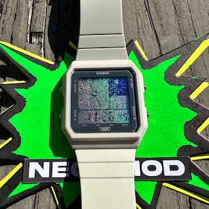Pu&ograve; includere: Un orologio digitale Casio grigio chiaro con quadrante quadrato e schermo olografico. L'orologio mostra l'ora digitale, il testo CASIO e WR, e pulsanti laterali etichettati come ADJUST, LIGHT, MODE e SEARCH. Il cinturino &egrave; grigio chiaro.