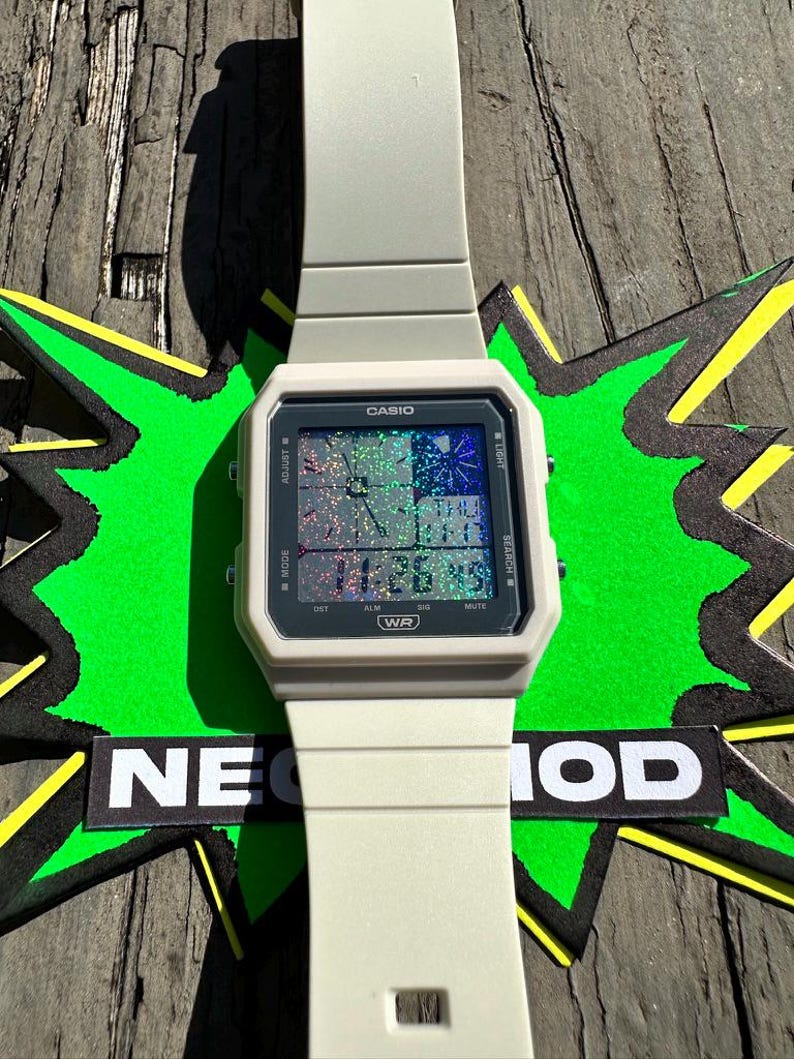 Pu&ograve; includere: Un orologio digitale Casio grigio chiaro con quadrante quadrato e schermo olografico. L'orologio mostra l'ora digitale, il testo CASIO e WR, e pulsanti laterali etichettati come ADJUST, LIGHT, MODE e SEARCH. Il cinturino &egrave; grigio chiaro.