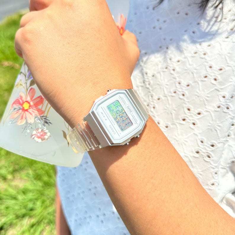 May include: A digital wristwatch with a silver-toned rectangular case and a clear, transparent resin strap. The watch face features a digital display with the Casio brand name, the model designation F-91W, and the letters WR printed on the front.