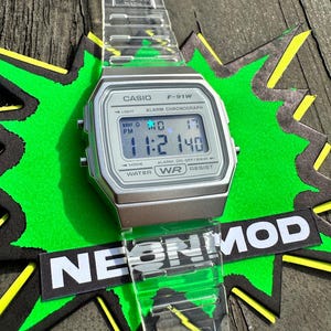 May include: A Casio F-91W digital watch with a clear, transparent resin strap and a silver-toned rectangular case. The digital display shows the time 11:21 and the date MO 17. The watch face features the text CASIO, F-91W, ALARM CHRONOGRAPH, and WATER WR RESIST.