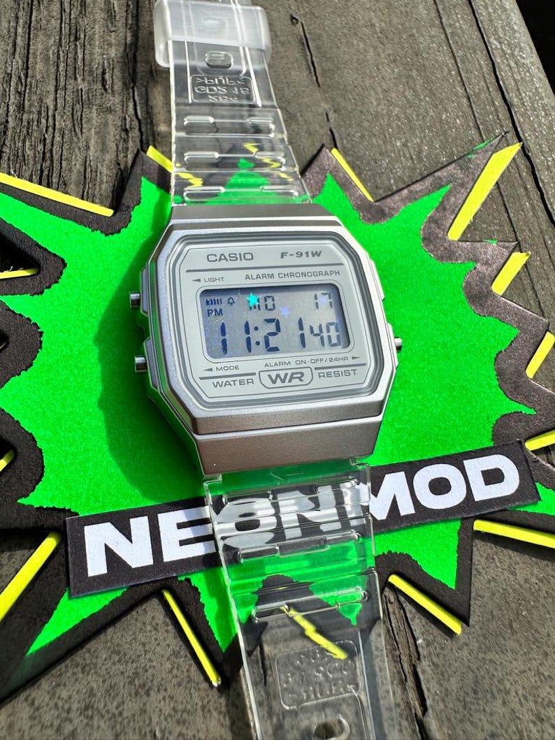 May include: A Casio F-91W digital watch with a clear, transparent resin strap and a silver-toned rectangular case. The digital display shows the time 11:21 and the date MO 17. The watch face features the text CASIO, F-91W, ALARM CHRONOGRAPH, and WATER WR RESIST.
