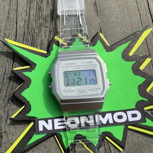 May include: A digital wristwatch with a silver-toned rectangular case and a clear, transparent resin strap. The watch face displays the text CASIO F-91W, ALARM CHRONOGRAPH, and WATER WR RESIST. The digital screen shows a holographic glitter effect.
