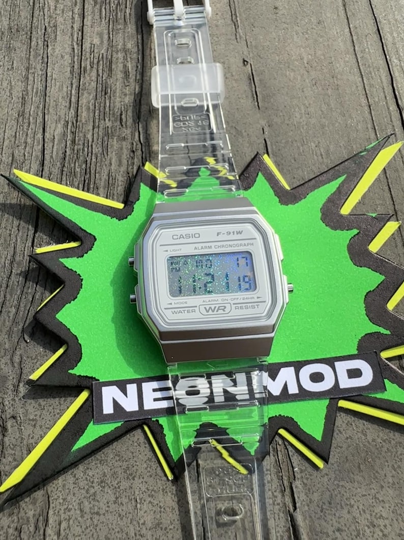 May include: A digital wristwatch with a silver-toned rectangular case and a clear, transparent resin strap. The watch face displays the text CASIO F-91W, ALARM CHRONOGRAPH, and WATER WR RESIST. The digital screen shows a holographic glitter effect.