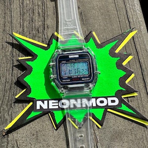 May include: A digital wristwatch with a clear, transparent resin strap and a square case. The watch face features the brand name SKMEI, the text ALARM CHRONOGRAPH, and WATER 50M RESIST. The digital display shows the time 11:16 with a glittery effect.