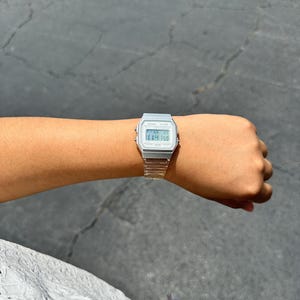 May include: A silver-toned digital wristwatch with a rectangular face and a clear, transparent resin strap. The watch features a digital display showing the time, date, and day of the week, with small buttons on the sides of the metallic-colored case.