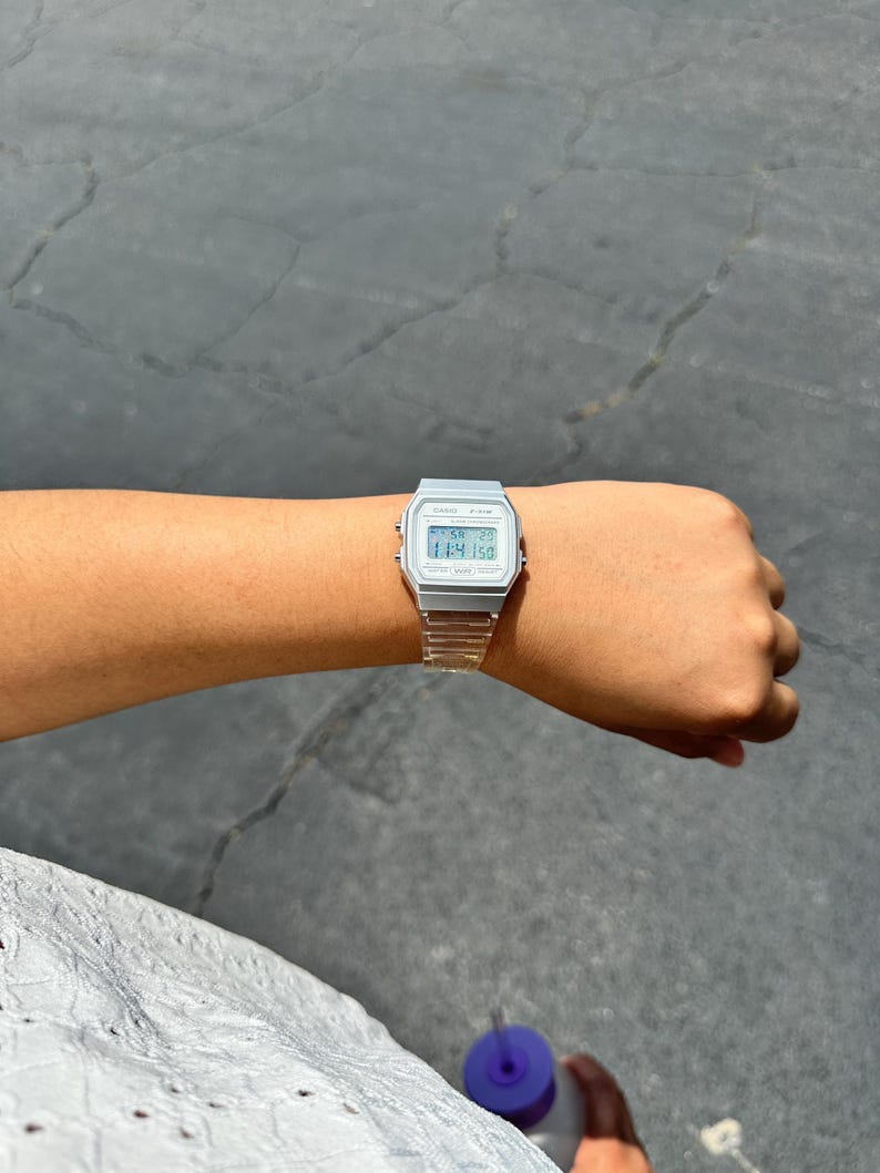 May include: A silver-toned digital wristwatch with a rectangular face and a clear, transparent resin strap. The watch features a digital display showing the time, date, and day of the week, with small buttons on the sides of the metallic-colored case.