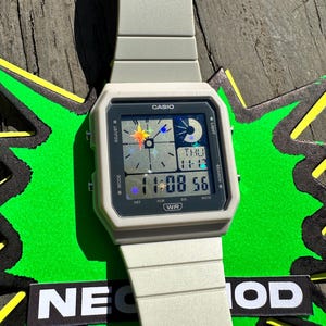 Pu&ograve; includere: Un orologio da polso digitale color beige chiaro con cassa quadrata e cinturino in resina abbinato. Il quadrante presenta il logo Casio, un display digitale che mostra 11:08 56 e grafiche olografiche di stelle e lune sullo schermo.