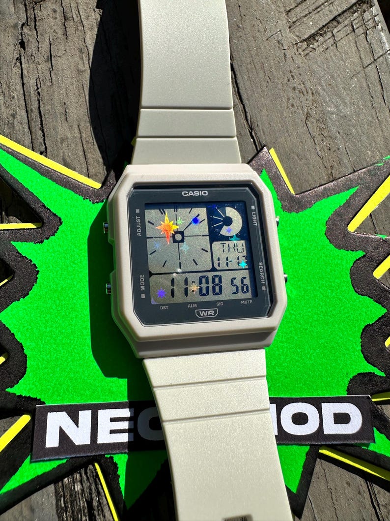 Pu&ograve; includere: Un orologio da polso digitale color beige chiaro con cassa quadrata e cinturino in resina abbinato. Il quadrante presenta il logo Casio, un display digitale che mostra 11:08 56 e grafiche olografiche di stelle e lune sullo schermo.