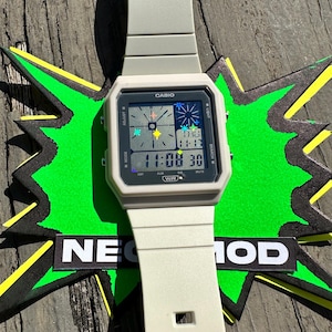 Pu&ograve; includere: Un orologio digitale Casio grigio chiaro con quadrante quadrato e cinturino in resina abbinato. Il display presenta una grafica a stella multicolore, l'ora 11:08, la data e il testo WR. L'orologio &egrave; posizionato su una forma a stella verde con il testo NEO MOD.