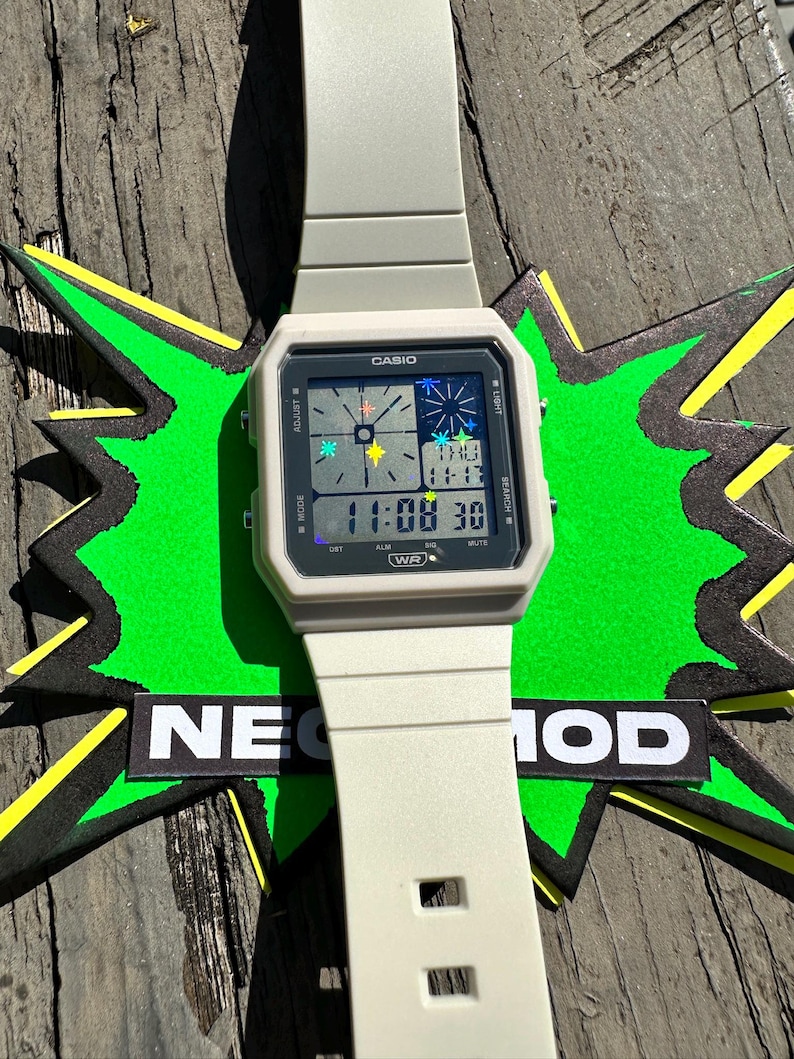 Pu&ograve; includere: Un orologio digitale Casio grigio chiaro con quadrante quadrato e cinturino in resina abbinato. Il display presenta una grafica a stella multicolore, l'ora 11:08, la data e il testo WR. L'orologio &egrave; posizionato su una forma a stella verde con il testo NEO MOD.