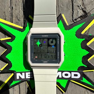 Pu&ograve; includere: Un orologio da polso digitale color bianco sporco opaco con cassa quadrata e cinturino in resina abbinato. Il quadrante presenta il logo Casio, un display digitale con icone a forma di stella e la scritta WR. Mostra l'ora 11:04 e il giorno THU.