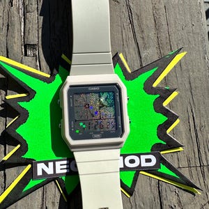 May include: A light beige Casio digital watch with a rectangular face and a matching silicone strap. The watch display features a holographic, glittery background with small green star icons and the text CASIO and WR printed on the frame.