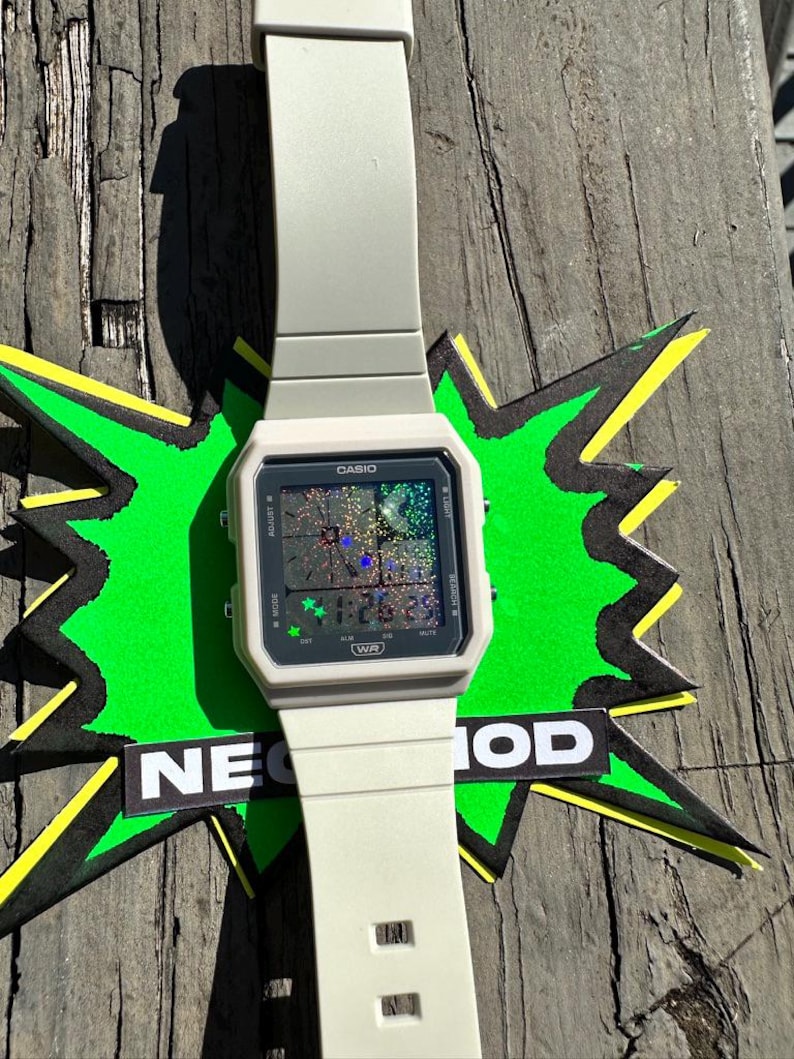 Pu&ograve; includere: Un orologio digitale Casio color beige chiaro con quadrante rettangolare e cinturino in silicone abbinato. Il display dell'orologio presenta uno sfondo olografico scintillante con piccole icone a stella verdi e il testo CASIO e WR.