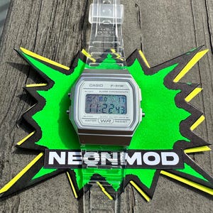 May include: A Casio F-91W digital watch with a silver-toned case and a clear, transparent resin strap. The watch face features a holographic display showing the time 11:22:43. The watch is positioned over a green and black starburst graphic with the text NEONMOD.