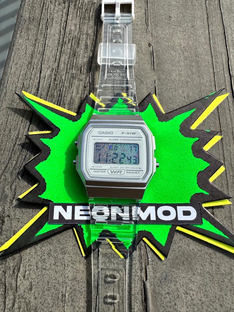 May include: A Casio F-91W digital watch with a silver-toned case and a clear, transparent resin strap. The watch face features a holographic display showing the time 11:22:43. The watch is positioned over a green and black starburst graphic with the text NEONMOD.