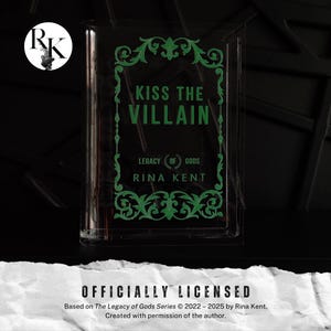 Könnte beinhalten: Ein durchsichtiges, rechteckiges Etui mit dem Text "KISS THE VILLAIN" in Grün. Das Etui zeigt auch den Text "LEGACY OF GODS" und "RINA KENT". Die Worte "OFFICIALLY LICENSED" stehen unten.