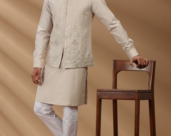May include: A man wearing a beige kurta and a beige waistcoat with silver embroidery. He is wearing white pants and brown sandals. He is standing next to a wooden chair.