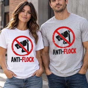 Może przedstawiać: Biały t-shirt i szary t-shirt, każdy z grafiką czarnej kamery monitoringu wewnątrz czerwonego koła zakazu z przekreśleniem. Poniżej grafiki znajduje się napis ANTI-FLOCK wykonany czarnymi i czerwonymi wielkimi literami.