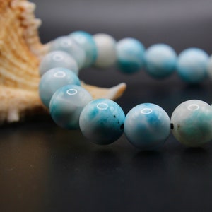 Puede incluir: Un brazalete de piedras preciosas azules y blancas con cuentas redondas. El brazalete está hecho de piedras naturales y está encordado en un cordón negro.