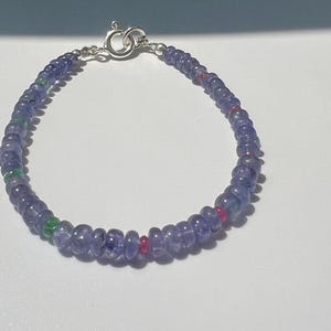 Puede incluir: Una pulsera de cuentas con pequeñas cuentas redondas y translúcidas de tanzanita azul bígaro, intercaladas con cuentas de acento ocasionales en verde vibrante y rojo brillante, con un cierre de resorte de plata pulida.