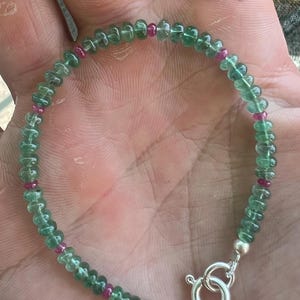 Può includere: Un braccialetto di perline verdi e rosa con una chiusura argentata. Il braccialetto è fatto di piccole perline rotonde che sono infilate su una catena d'argento.