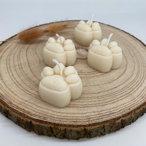 May include: Four white paw print shaped candles on a wooden slice. The candles are made of wax and have a white wick.