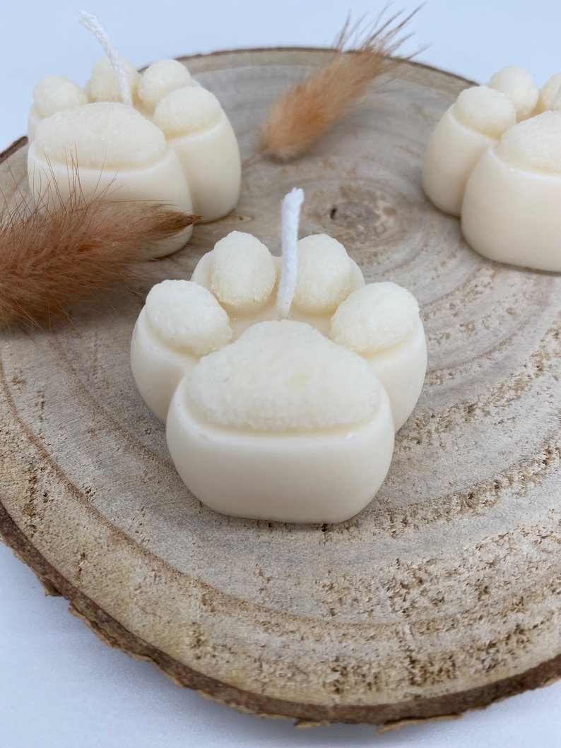 May include: Three white paw print shaped candles on a wooden slice. Each candle has a white wick.