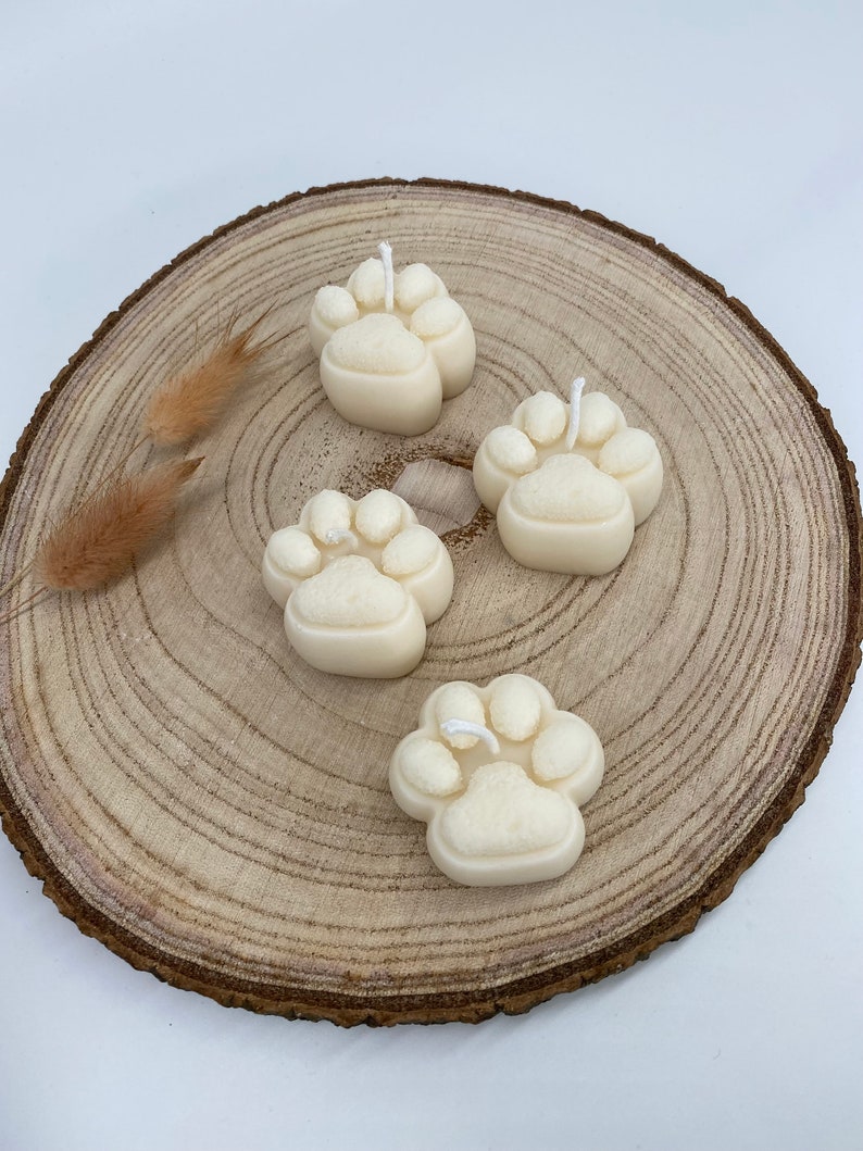 May include: Four white paw print candles on a wooden slice. The candles are made of a textured wax and have a white wick.