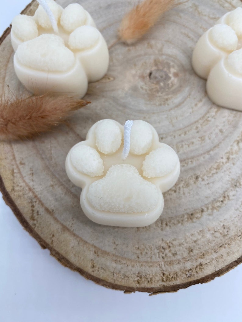 May include: Three white paw print shaped candles on a wooden surface. The candles have a white wick in the center.