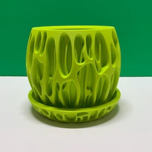 Pode incluir: Vaso de planta verde brilhante com pires a condizer. O vaso tem um design aberto e orgânico com numerosas aberturas ovais. O vaso e o pires são feitos de um material verde limão sólido. O diâmetro é de cerca de 15 cm.
