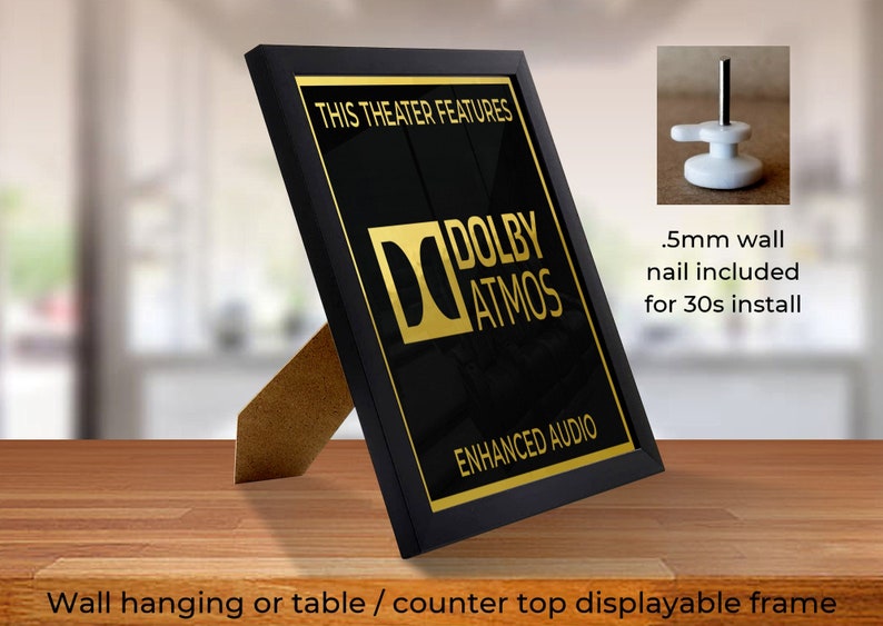 May include: Black framed sign with gold lettering that reads "This Theater Features Dolby Atmos Enhanced Audio". The sign is on a wooden table and can be hung on a wall.