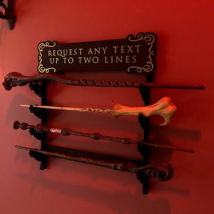 May include: A wooden wall-mounted wand holder with a sign that reads "REQUEST ANY TEXT UP TO TWO LINES". The holder has four wands displayed on it.