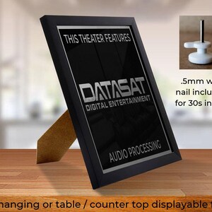 May include: A black framed sign with the text "THIS THEATER FEATURES DATASAT DIGITAL ENTERTAINMENT AUDIO PROCESSING". The sign is displayed on a wooden surface and includes a small wall nail for installation.