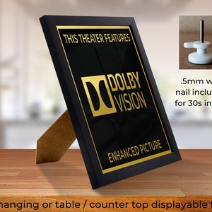 May include: Black framed picture with a gold Dolby Vision logo and the text "This Theater Features Enhanced Picture". The frame has a stand for tabletop display and a nail for wall hanging.