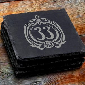 May include: A set of black slate coasters with a gray engraved design featuring a stylized castle icon and the number 33.