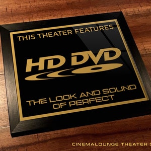 May include: Black framed sign with gold text that reads "This theater features HD DVD - The look and sound of perfect."  The sign also includes the HD DVD logo.  The sign is on a wood background.  Cinemalounge Theater Signs.