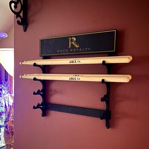 May include: A black metal drumstick holder mounted on a burgundy wall. The holder has two levels to hold drumsticks. The top level has a sign that reads "ROCK ROYALTY". Two drumsticks are in the holder, each with "ARLX 5A" printed on them.