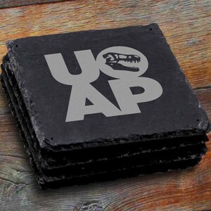 May include: A set of four black slate coasters with a dinosaur skull graphic and the text "U.O.A.P." in silver lettering.