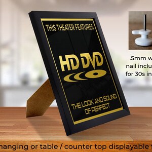 May include: Black framed picture with a gold inscription that reads "This theater features HD DVD The look and sound of perfect". The frame is standing on a wooden surface. A close-up of a white wall hook with a nail is shown in the upper right corner of the image.