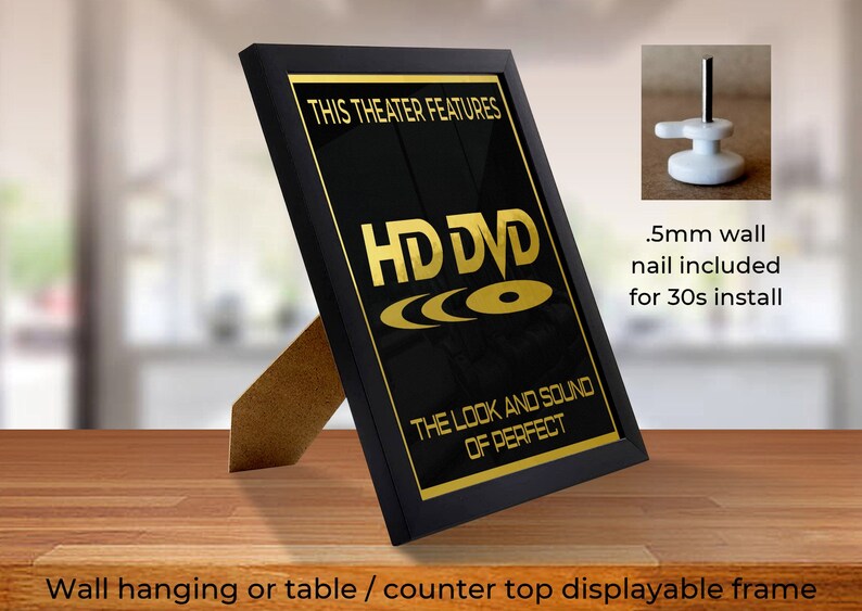 May include: Black framed picture with a gold inscription that reads "This theater features HD DVD The look and sound of perfect". The frame is standing on a wooden surface. A close-up of a white wall hook with a nail is shown in the upper right corner of the image.