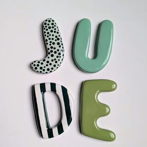 May include: Four ceramic letters spelling JUDE. The J is mint green with black polka dots. The U is a solid light mint green. The D features vertical dark forest green stripes on a white base. The E is a solid muted olive green colour.