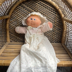 May include: A doll with a round peach-colored face, blue eyes, and a soft vinyl head. The doll wears a cream-colored bonnet with lace trim and a matching long-sleeved dress featuring a delicate pink leaf pattern, white lace accents, and a bow.