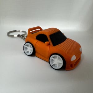 May include: A 3D-printed orange sports car keychain featuring a compact, stylized design with a rear spoiler, black windows, and white five-spoke wheels. The car body has a visible layered texture, and a silver metal split ring is attached to the rear.