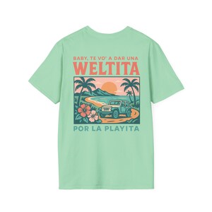 May include: A light green t-shirt featuring a beach scene with a blue jeep, palm trees, and flowers. The text on the shirt reads "BABY, TE VO' A DAR UNA WELTITA POR LA PLAYITA" in orange and green.