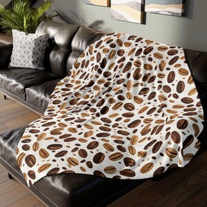 May include: A white throw blanket with a brown coffee bean pattern. The blanket is draped over a black leather couch.