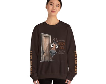 May include: A dark brown sweatshirt featuring a cartoon horse peeking out from behind a wooden door. The text "You say HORSE GIRL like it's a bad thing..." is printed on the front. The sleeves have the words "HORSE GIRL" printed vertically.
