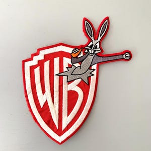 May include: A red and white embroidered patch featuring the Warner Bros. logo with Bugs Bunny holding a carrot and punching through the shield.