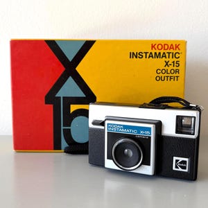 May include: A vintage Kodak Instamatic X-15 camera with its original box. The camera is black and white with a silver lens and a black strap. The box is red, yellow, and blue with the words "KODAK INSTAMATIC X-15 COLOR OUTFIT".