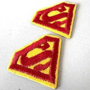 May include: Two embroidered patches featuring the iconic Superman logo. The patches have a yellow background with a red "S" emblem. The patches are likely for clothing or accessories.